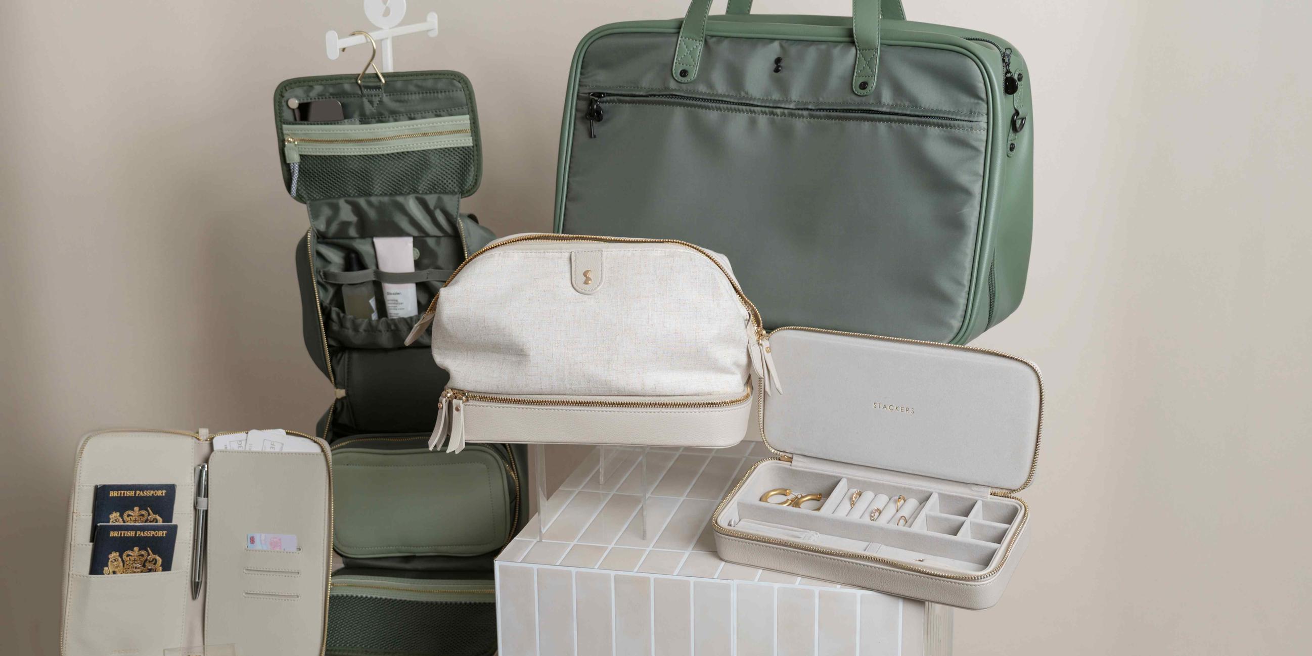 Stackers travel collection including a green hanging toiletry bag, green weekender holdall, cream linen wash bag, grey jewellery case, and passport travel wallet with British passports
